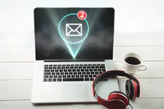 Laptop na biurku z otwartą pocztą internetową, obok kubek kawy i słuchawki