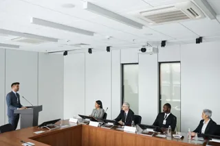 Narada przy stole konferencyjnym z mikrofonami i monitorami